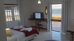 A room with a bed, a desk with a computer, a chair, a table, and a view of the beach. - Atlantico Hotel (Rio das Ostras)