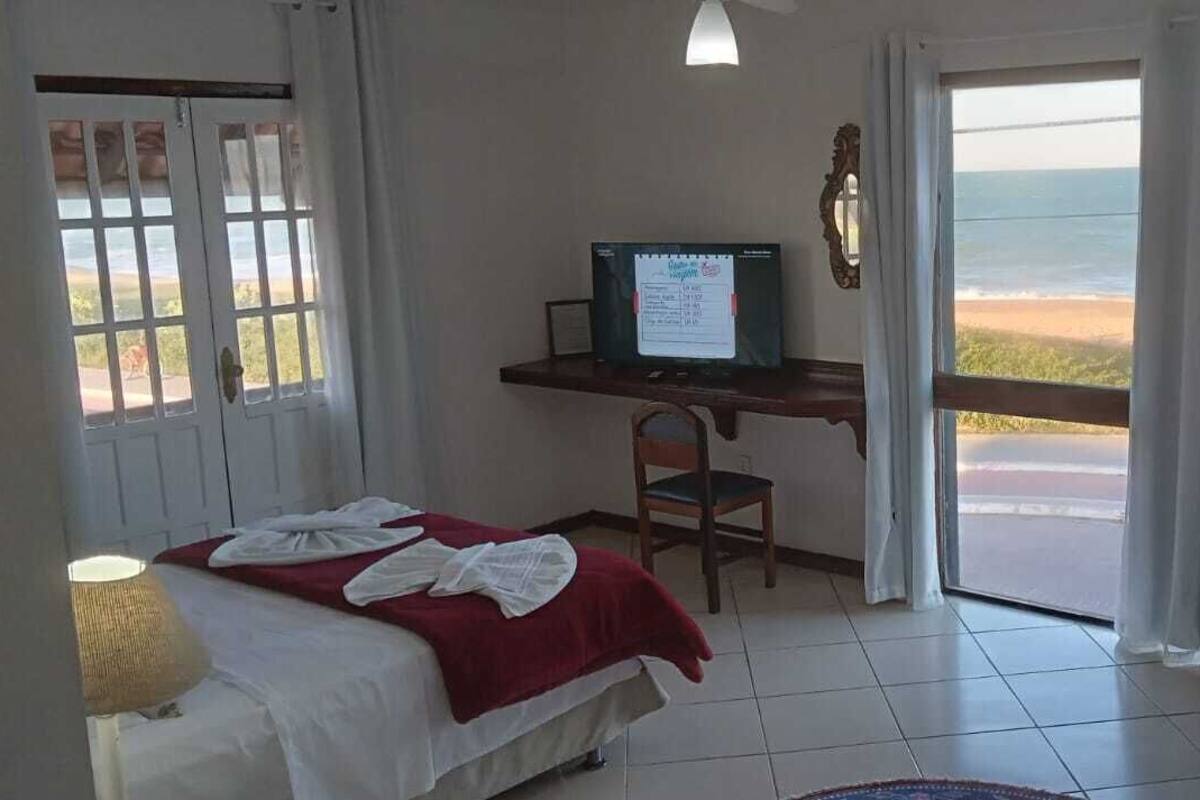 A room with a bed, a desk with a computer, a chair, a table, and a view of the beach.