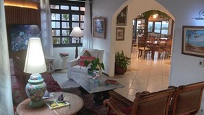 A cozy living area with a sofa, a coffee table, and a lamp. There is a dining area with a table and chairs visible through an archway. - Atlantico Hotel (Rio das Ostras)
