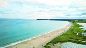 An aerial view of a beach with clear turquoise waters, a sandy shore, and a grassy area with a path leading to the beach.