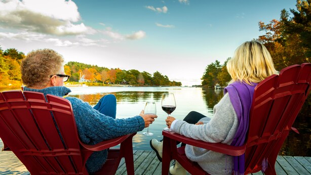Two people sitting on red chairs facing a lake with autumn foliage, holding wine glasses.
