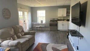 A living room with a beige sofa, a dining table with chairs, a kitchen area with cabinets and appliances, and a mounted television.