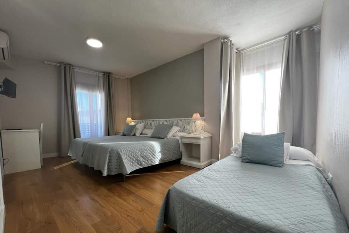 A hotel room with two beds, a bedside table with a lamp, a small desk, and a window with curtains.
