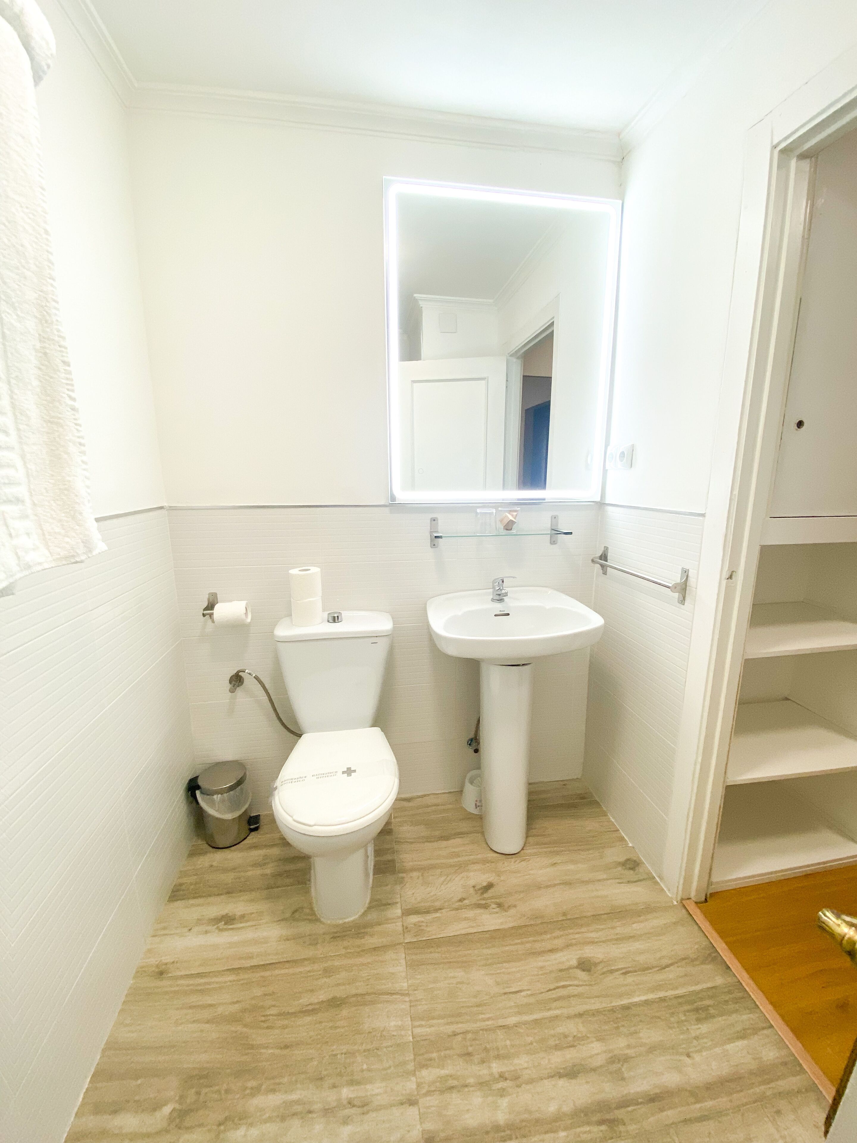 Baño pequeño y blanco con inodoro, lavabo y espejo. Hay un perchero en la pared y un bote de basura al lado del inodoro.