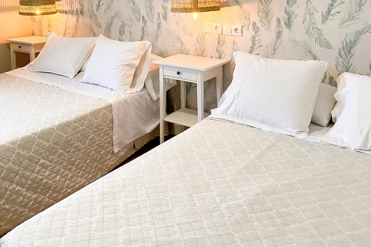 A hotel room with two beds, a small white nightstand, and a wall with a leafy pattern.