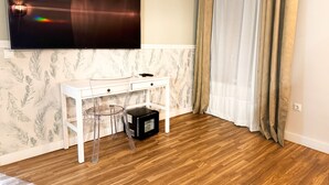 Een kamer met een wit bureau, een transparante glazen stoel, een wandgemonteerde flatscreen televisie en een houten vloer.