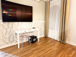 Una habitación con un escritorio blanco, una silla de vidrio transparente, un televisor de pantalla plana montado en la pared y piso de madera.