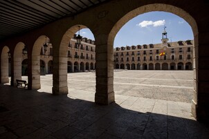 Un patio abierto con arcos de piedra y suelo de baldosas, que conduce a un edificio con múltiples ventanas y una bandera.