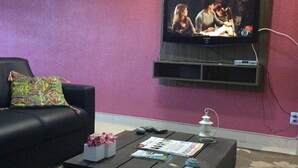 A living room with a black leather sofa, a coffee table with magazines and a decorative item, and a flat-screen TV mounted on a pink wall. - Hostel Le Monde (Angra dos Reis)