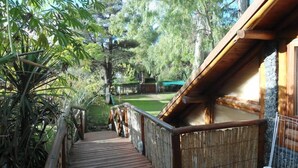 A wooden deck leading to a cabin with a stone wall and a view of a grassy yard and trees. - Cabañas Manamar (Villa Gesell)