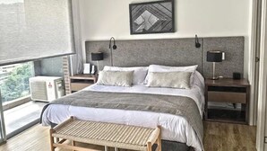 A modern bedroom with a large bed, two bedside tables with lamps, a bench, and a view of greenery through the window. - Urban Studios by Velvet Luxury (Medellín)