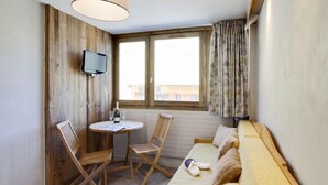 Una habitación acogedora con suelo de madera, una mesa redonda con sillas, un sofá, un televisor montado en la pared y una ventana con cortinas.
