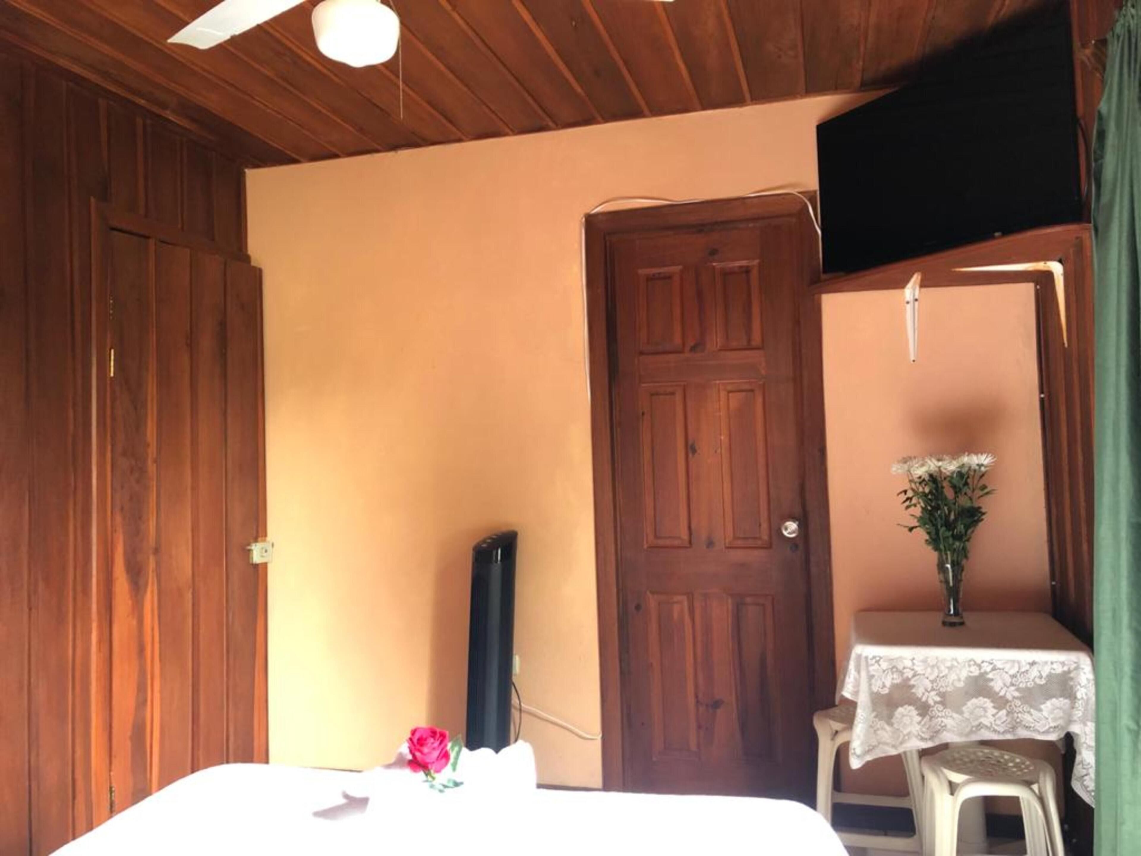 Une pièce avec un plafond en bois, un lit, une petite table sur laquelle est posé un vase de fleurs et une porte.