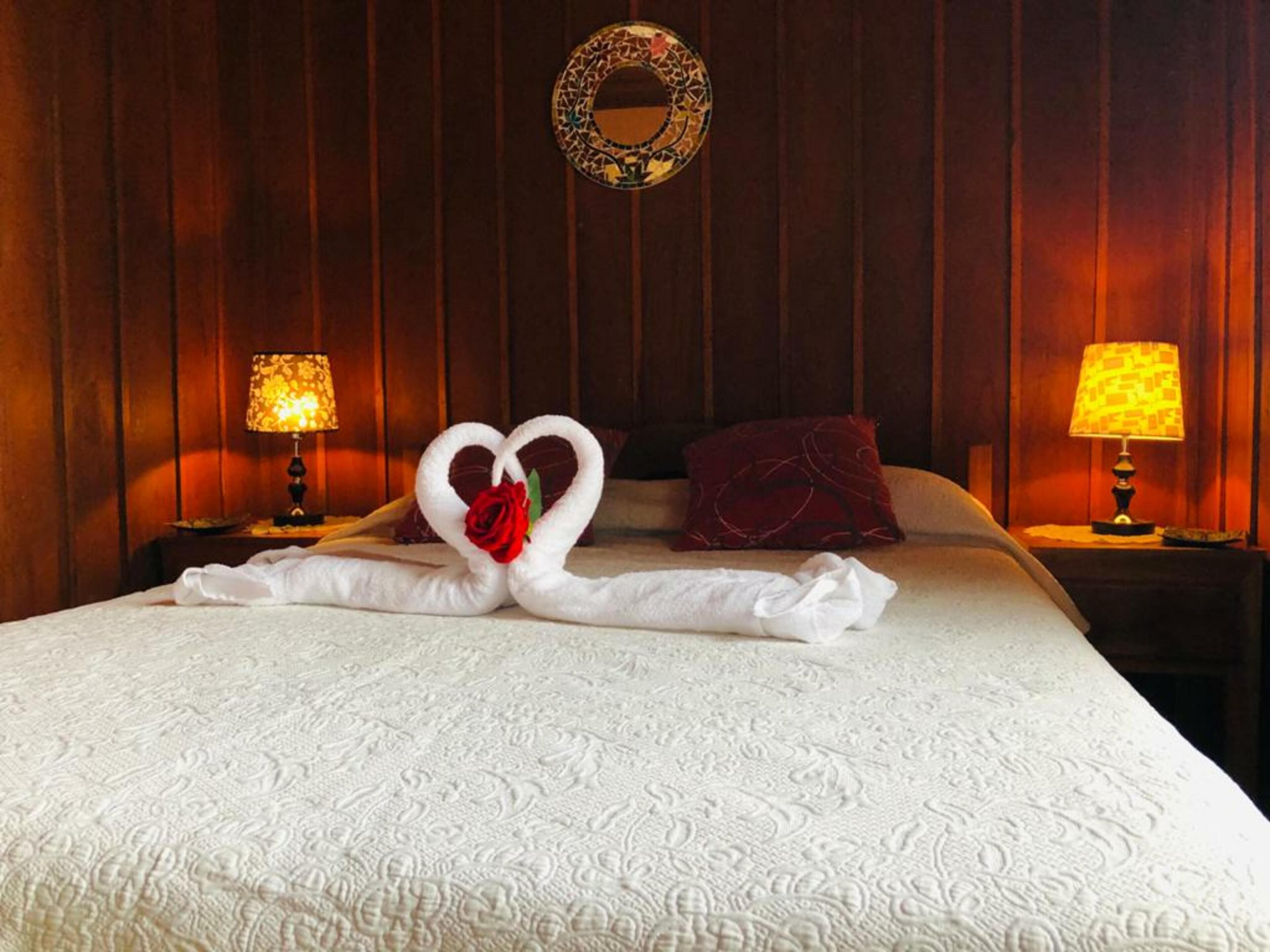 Een net opgemaakt bed met een handdoekarrangement in de vorm van een hart, inclusief een roos, twee lampen aan weerszijden en een ronde wanddecoratie.