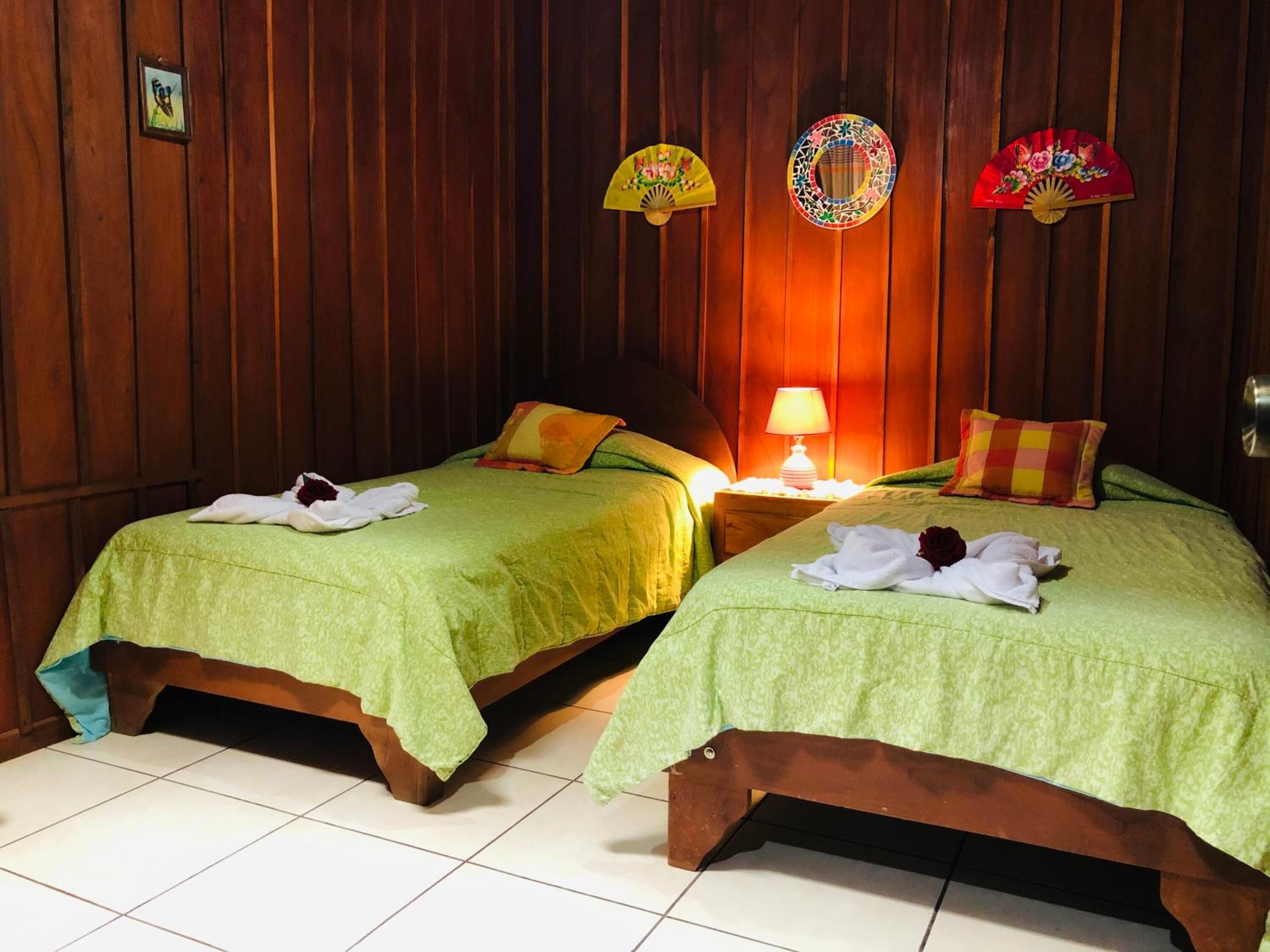 A room with two wooden beds, each with a green bedspread and pillows, a bedside table with a lamp, and decorative fans on the wall.