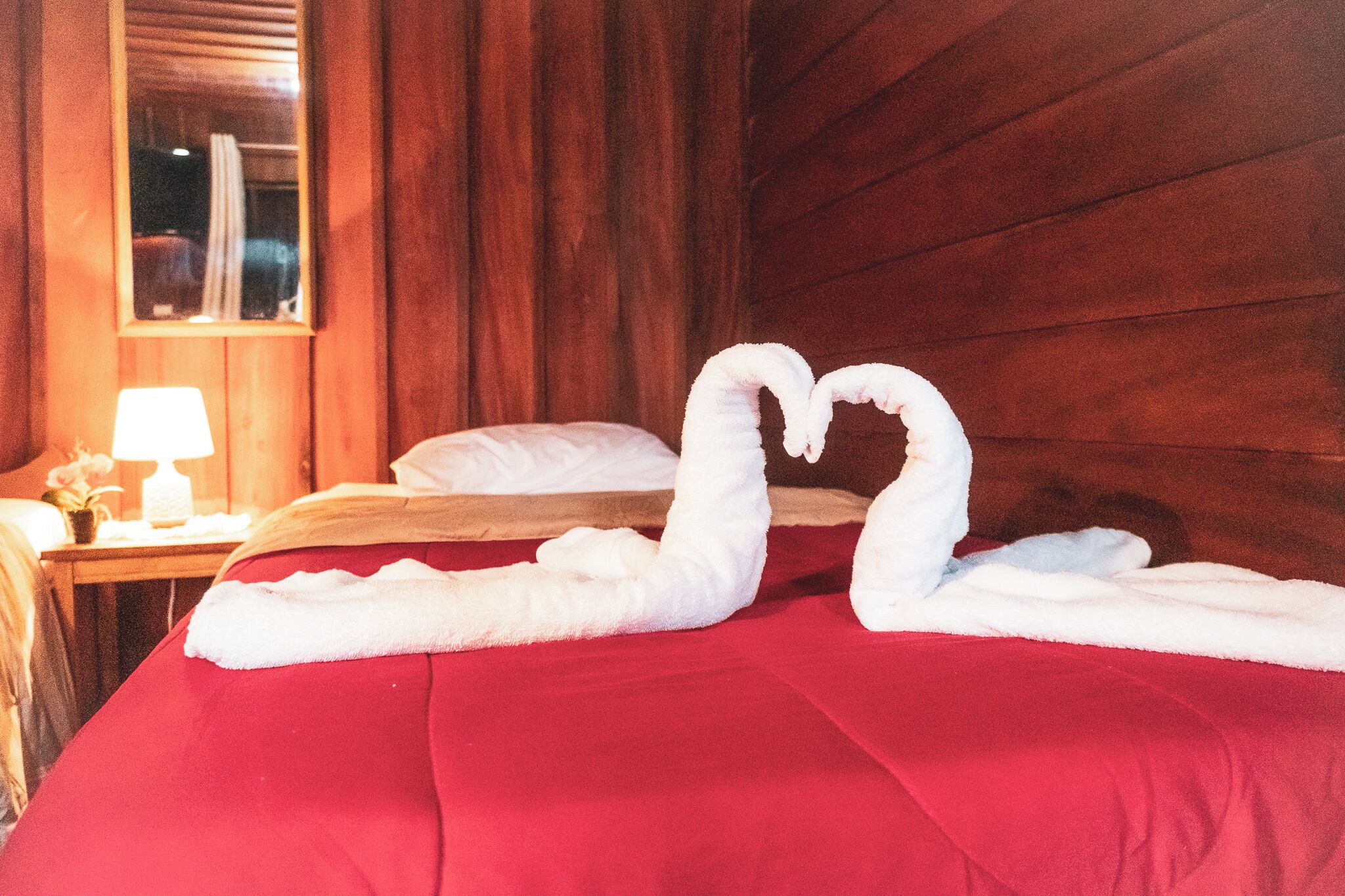 Une chambre d’hôtel avec une couvre-lit rouge, une tête de lit en bois, une table de chevet avec une lampe et une fenêtre avec des rideaux.