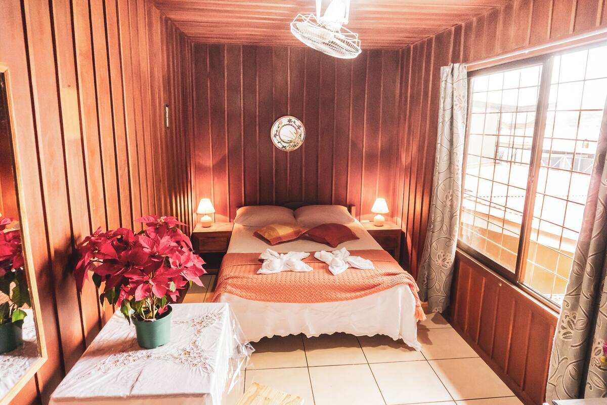A cozy room with wooden walls, a bed with pillows, a bedside table with a lamp, and a window with curtains.