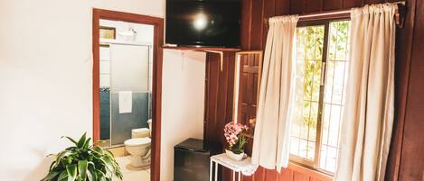 A room with a bed, a TV mounted on the wall, a small table with a flower vase, a potted plant, and a window with curtains.
