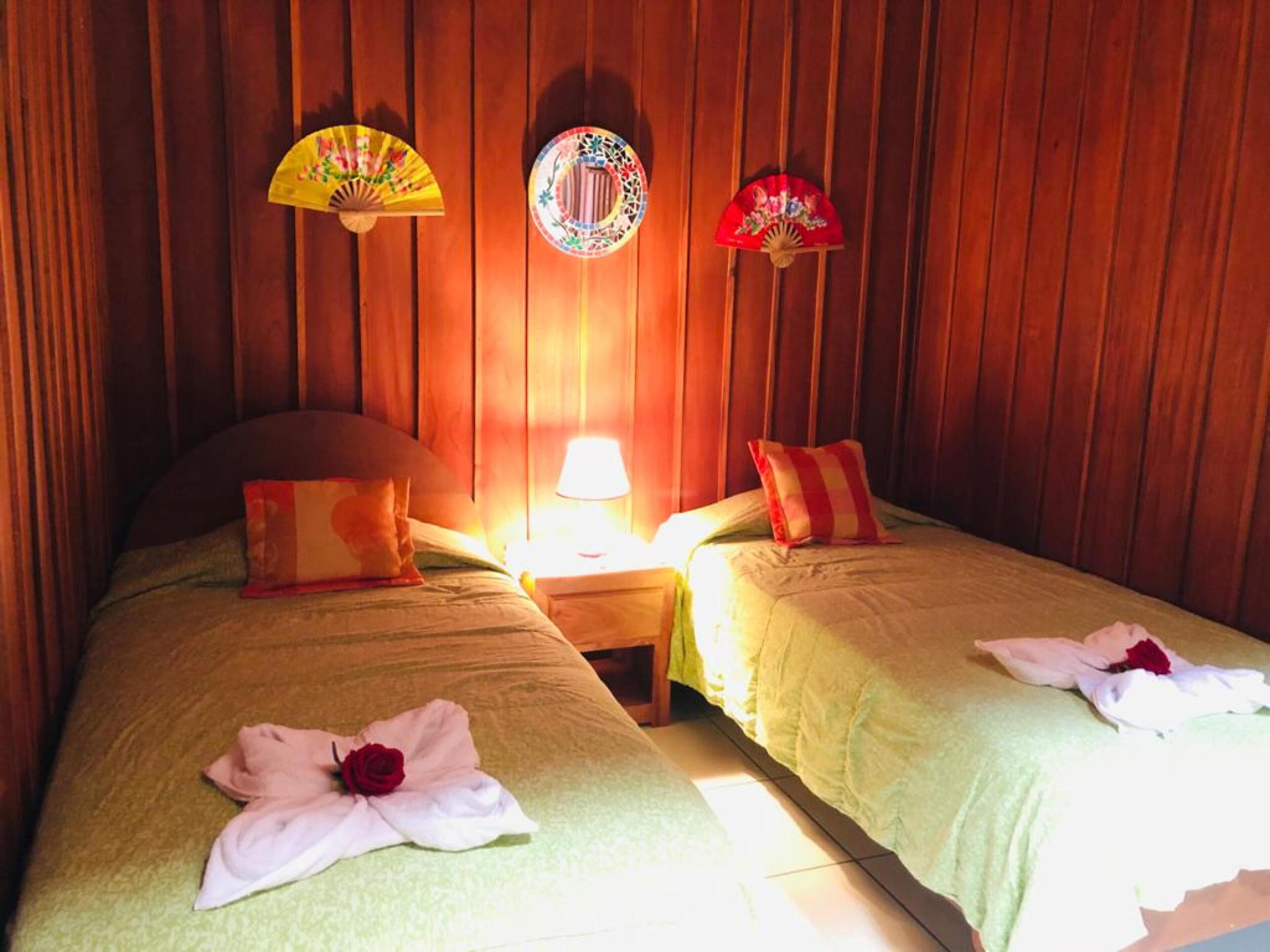 Quarto de hotel com duas camas, painéis de madeira, mesa de cabeceira com abajur e ventoinhas decorativas.