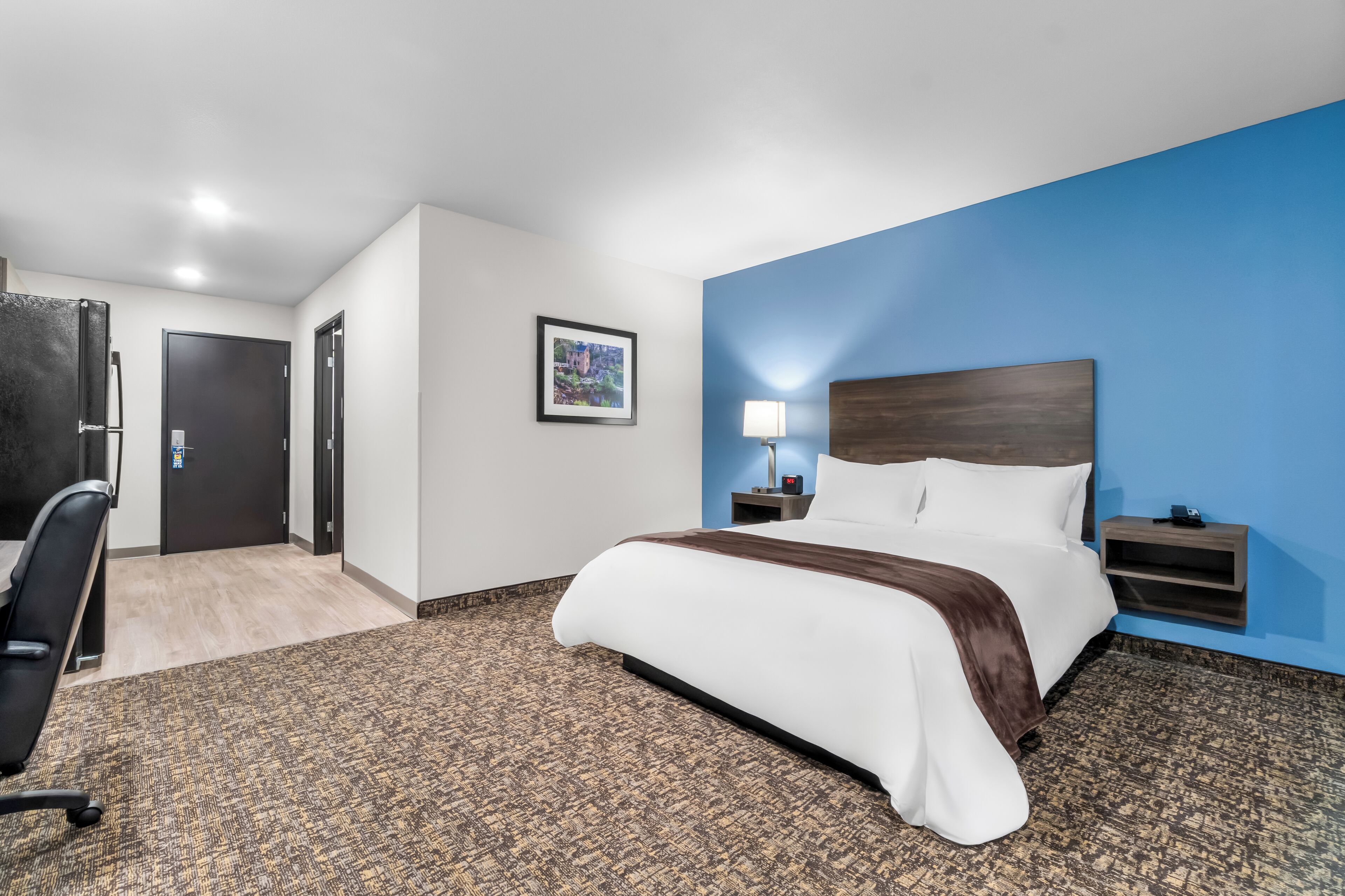 A hotel room with a bed, a desk, a chair, a nightstand, a lamp, and a picture on the wall.