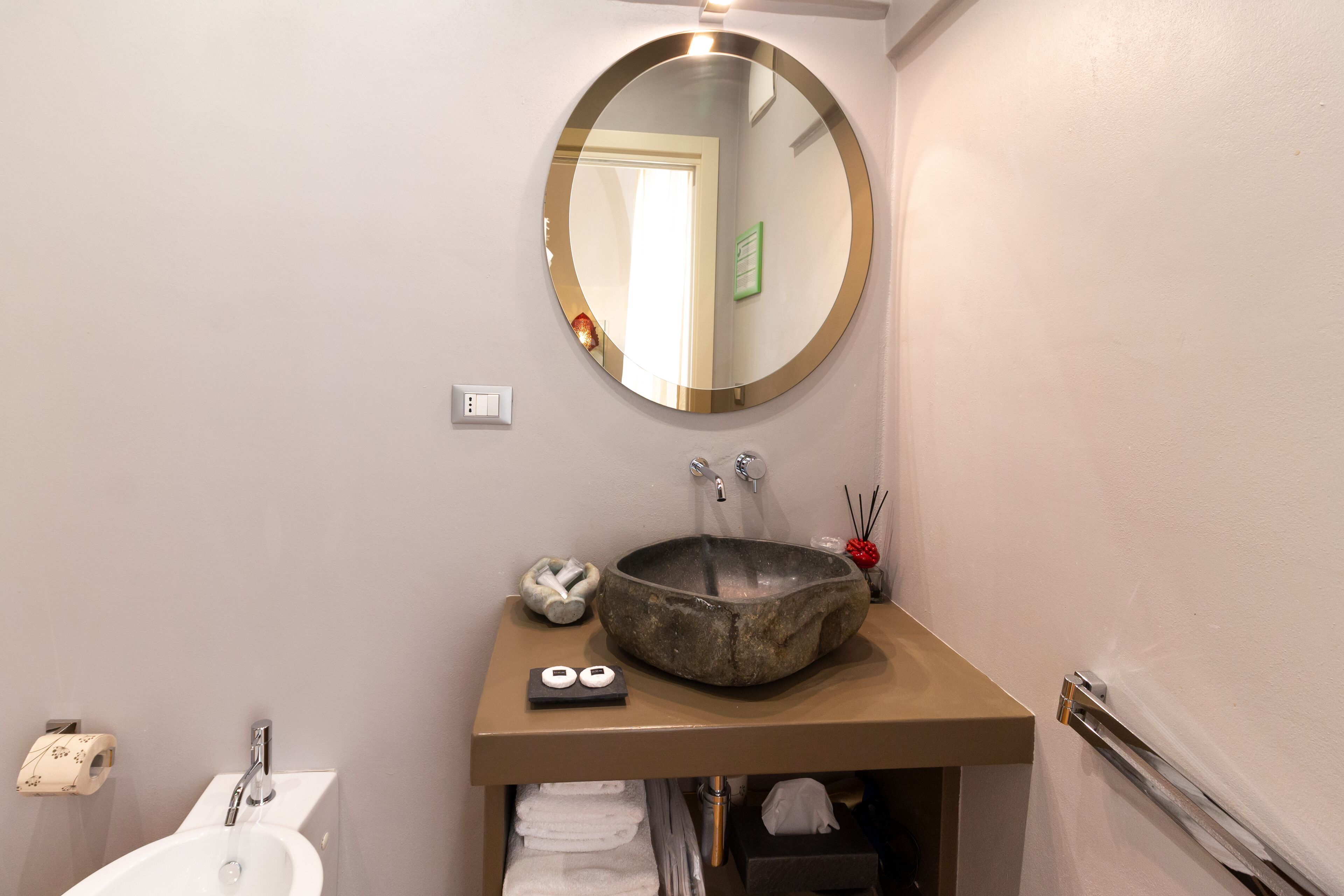 A bathroom with a round mirror, a stone sink, a towel rack, and a toilet.