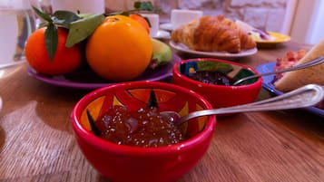 Una mesa de desayuno con frutas, cruasanes y un bol rojo de mermelada.