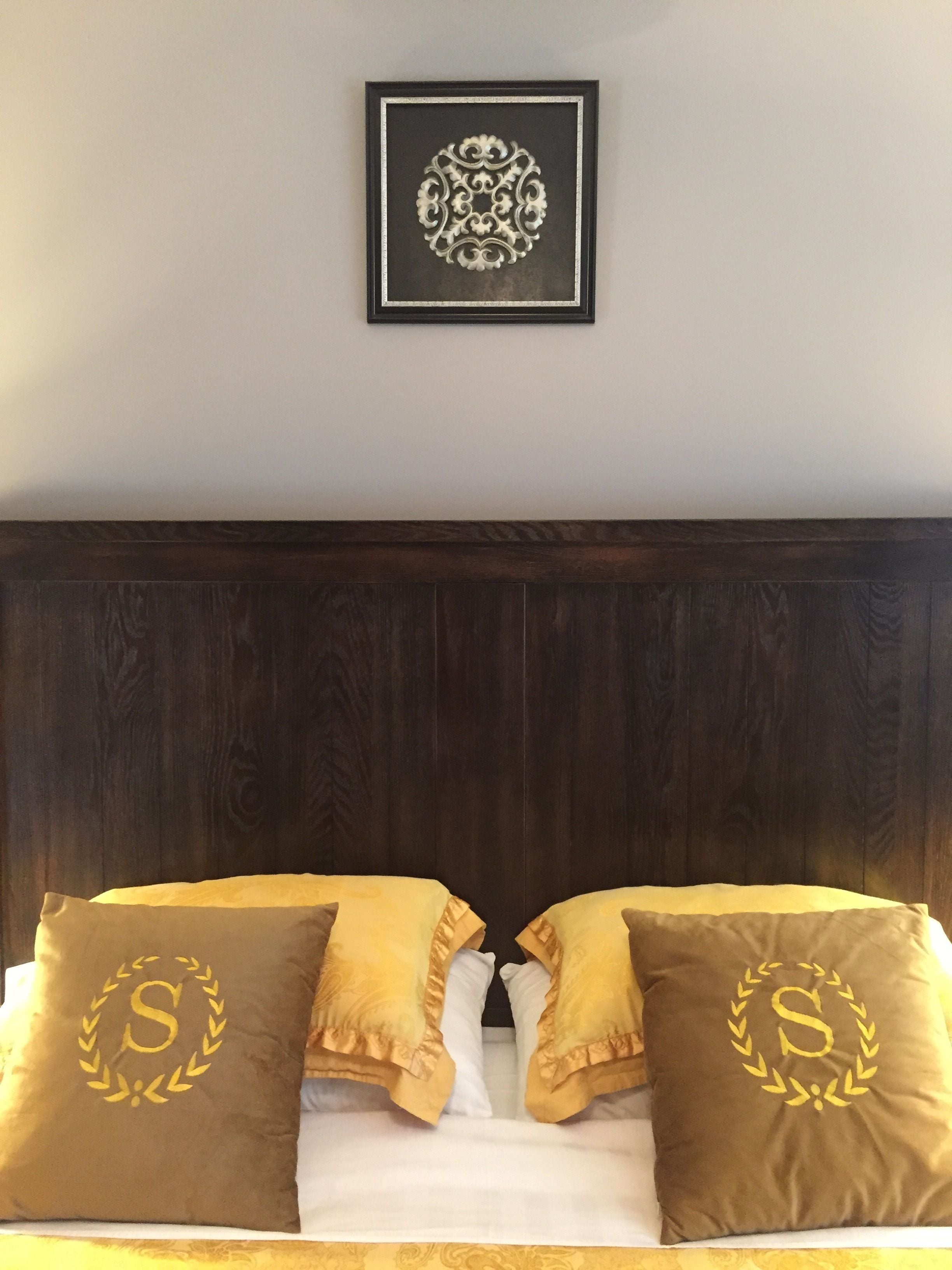 Un dormitorio con cabecera de madera oscura, una pieza decorativa enmarcada sobre la cama y dos almohadas con un monograma.