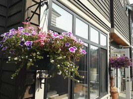 Uma cesta suspensa com flores roxas e brancas em frente a uma janela.