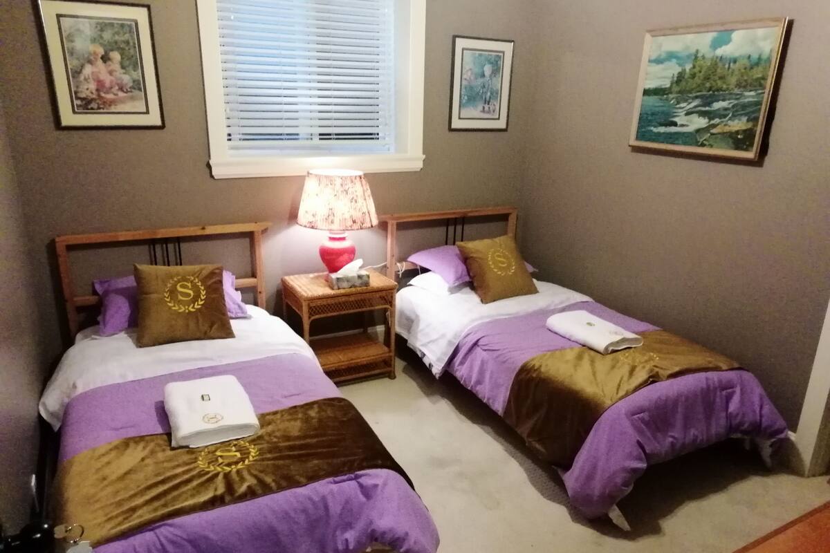 Ein Schlafzimmer mit zwei Betten, jeweils mit violett-brauner Bettwäsche, einem Nachttisch mit Lampe sowie gerahmten Bildern an den Wänden.