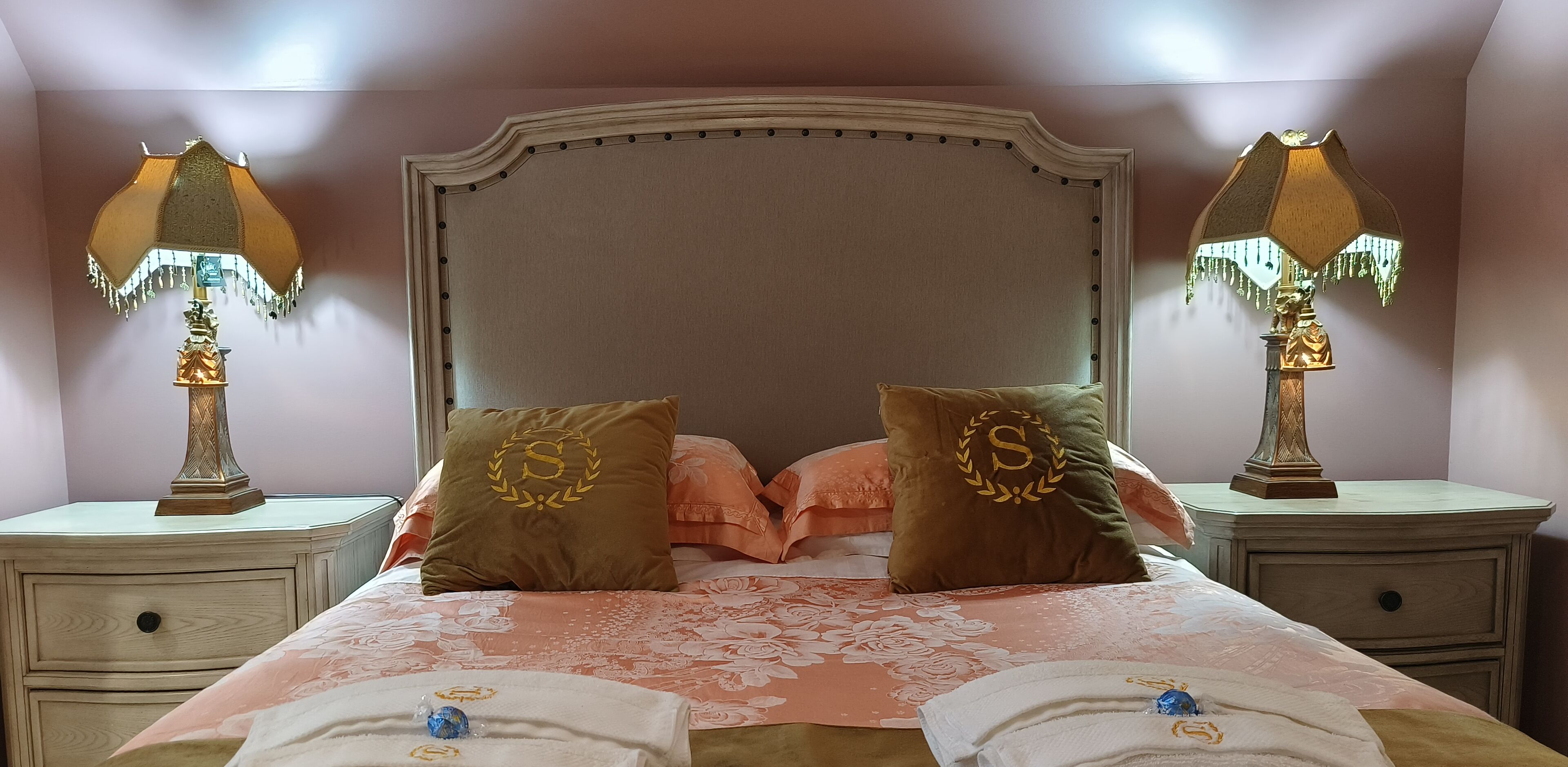Una cama bien hecha con ropa de cama floral, dos lámparas de noche y almohadas a juego con un monograma.