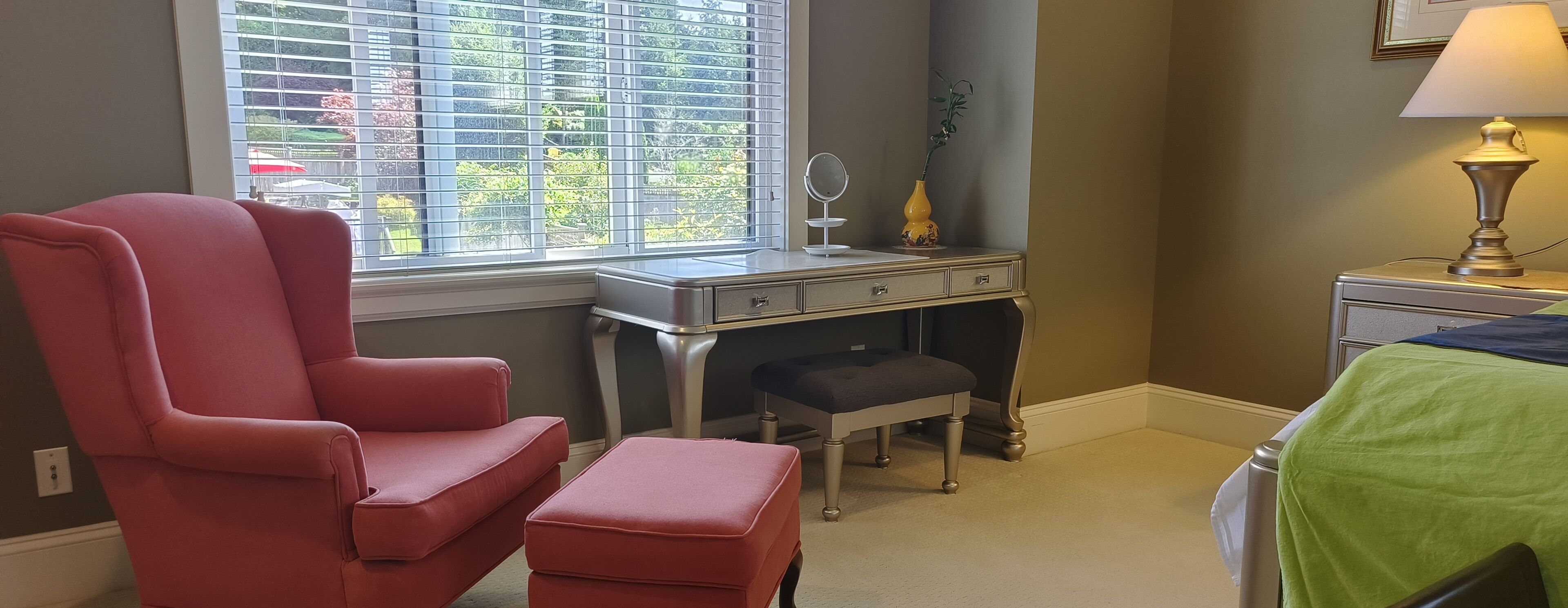A room with a red chair, a desk with drawers, a bench, a lamp, and a bed with a green cover.