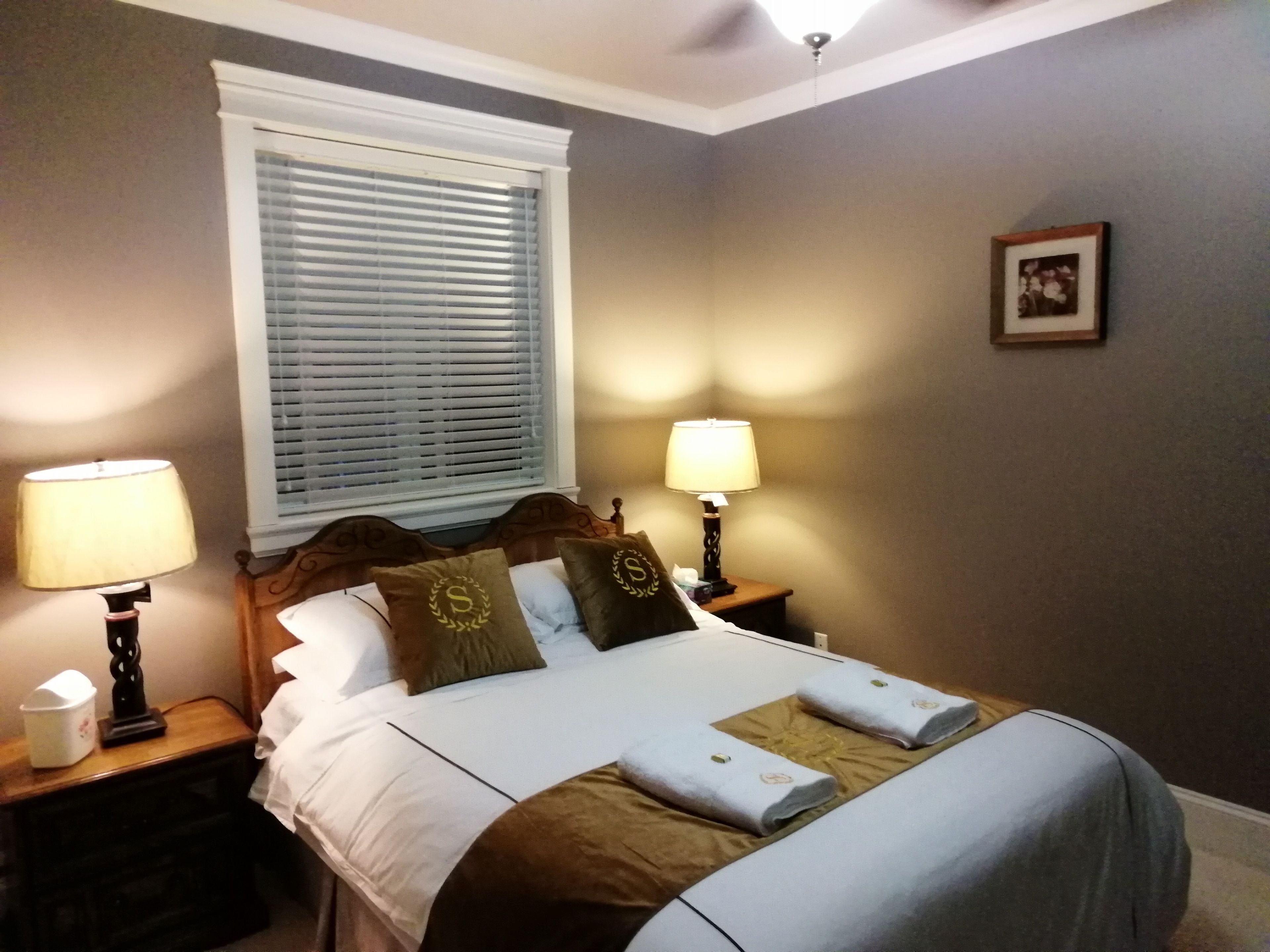 A neatly made bed with pillows and a blanket, two bedside lamps, a nightstand, a window with blinds, and a framed picture on the wall.