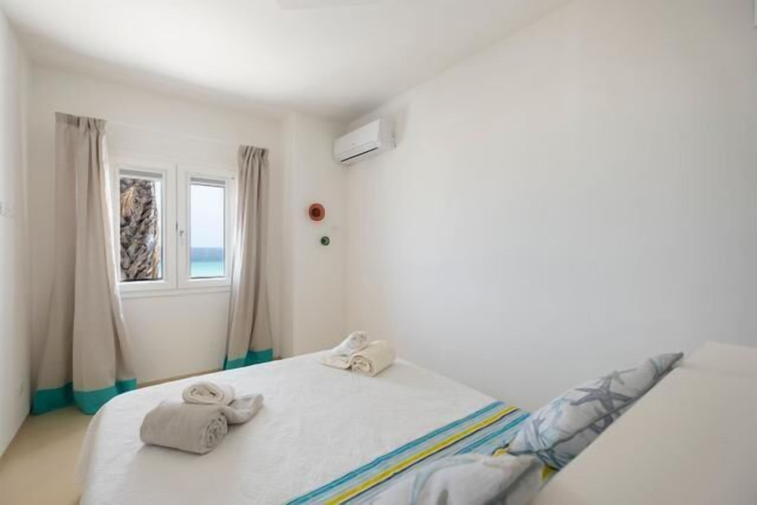 A hotel room with a bed, a window with curtains, an air conditioning unit, and a view of the sea.