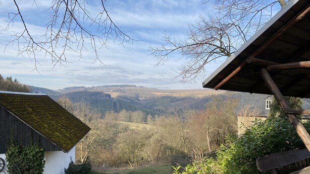 A rustic cabin with a sloped roof, a wooden bench, and a view of a valley with trees and hills.