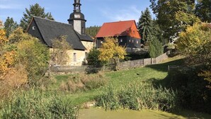 A traditional stone building with a red roof, a bell tower, and a wooden fence surrounding a grassy area with a pond.