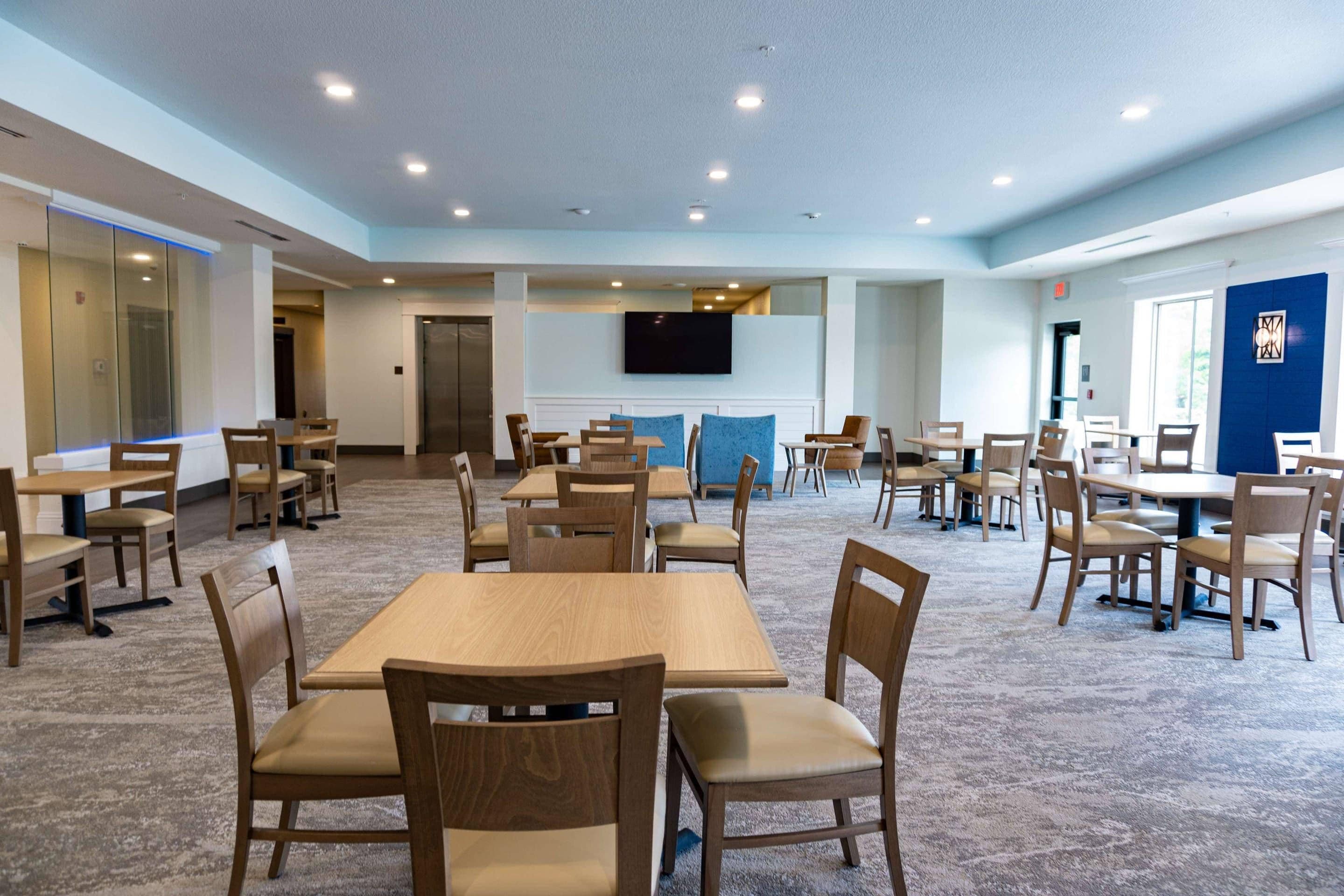 A spacious dining area with multiple tables and chairs, a flat-screen TV mounted on the wall, and a carpeted floor.
