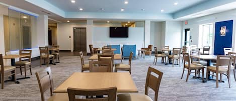 A spacious dining area with multiple tables and chairs, a flat-screen TV mounted on the wall, and a carpeted floor.