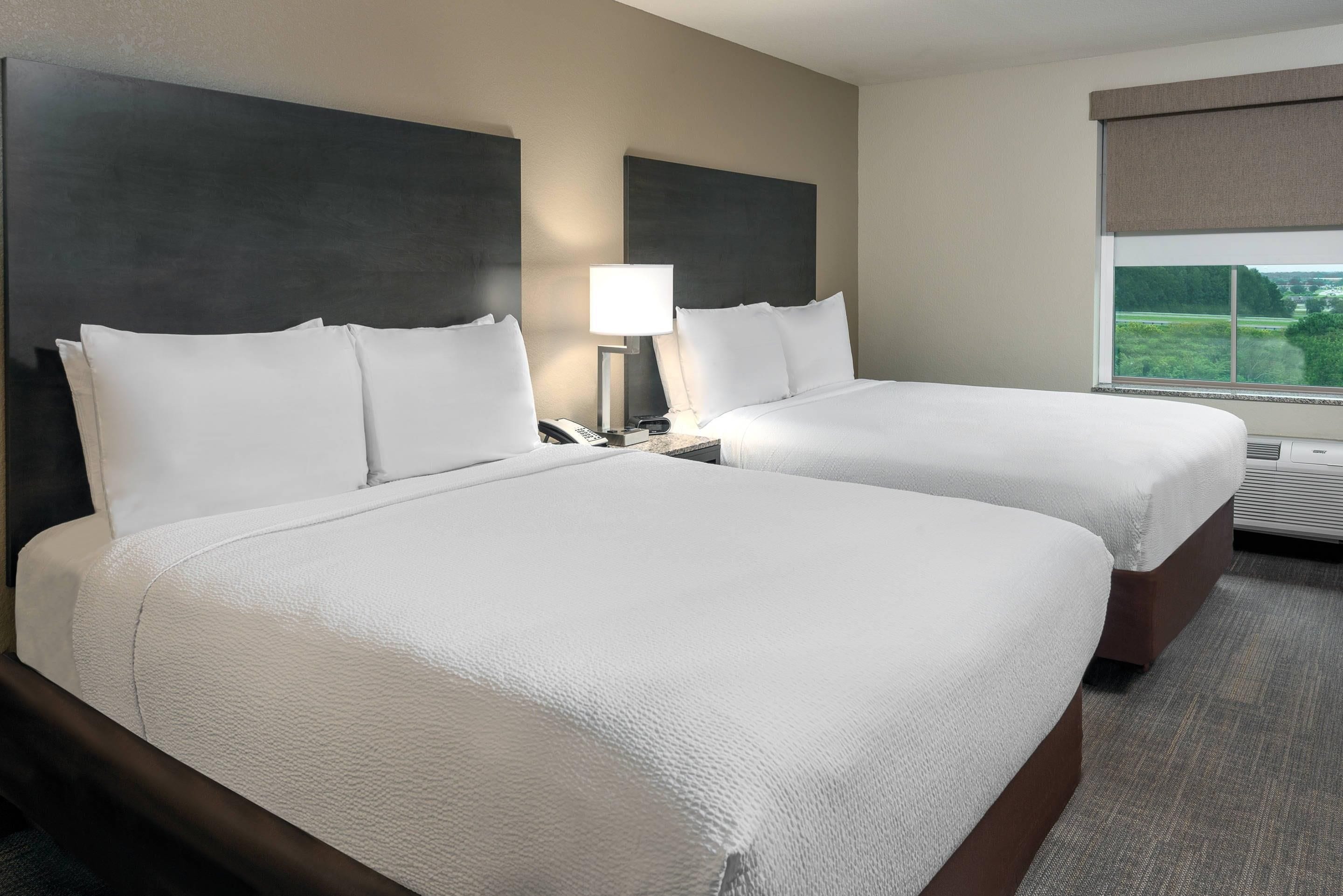 Een hotelkamer met twee bedden, een nachtkastje met lamp, een raam met uitzicht en een wandgemonteerde airconditioning.