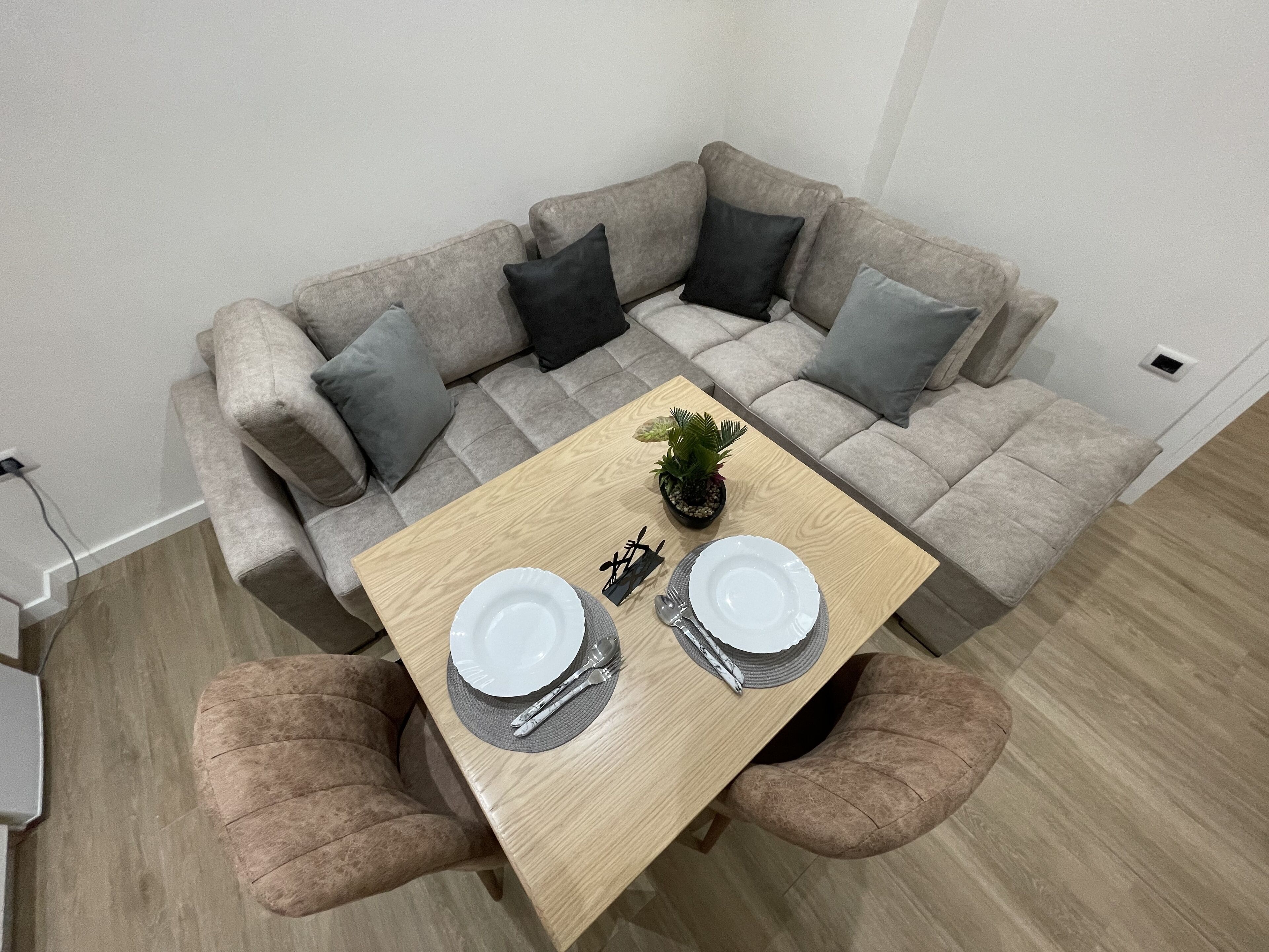 A dining area with a wooden table set for two, a grey sofa with cushions, and a small potted plant on the table.