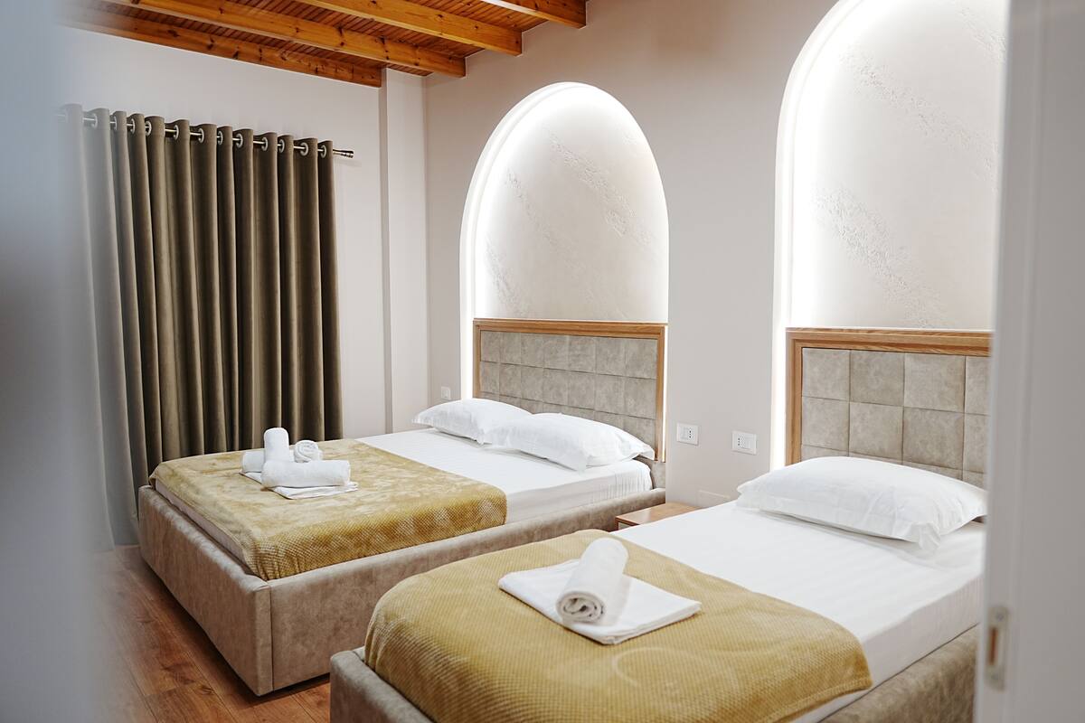 A hotel room with two beds, a wooden ceiling, and a large arched window.