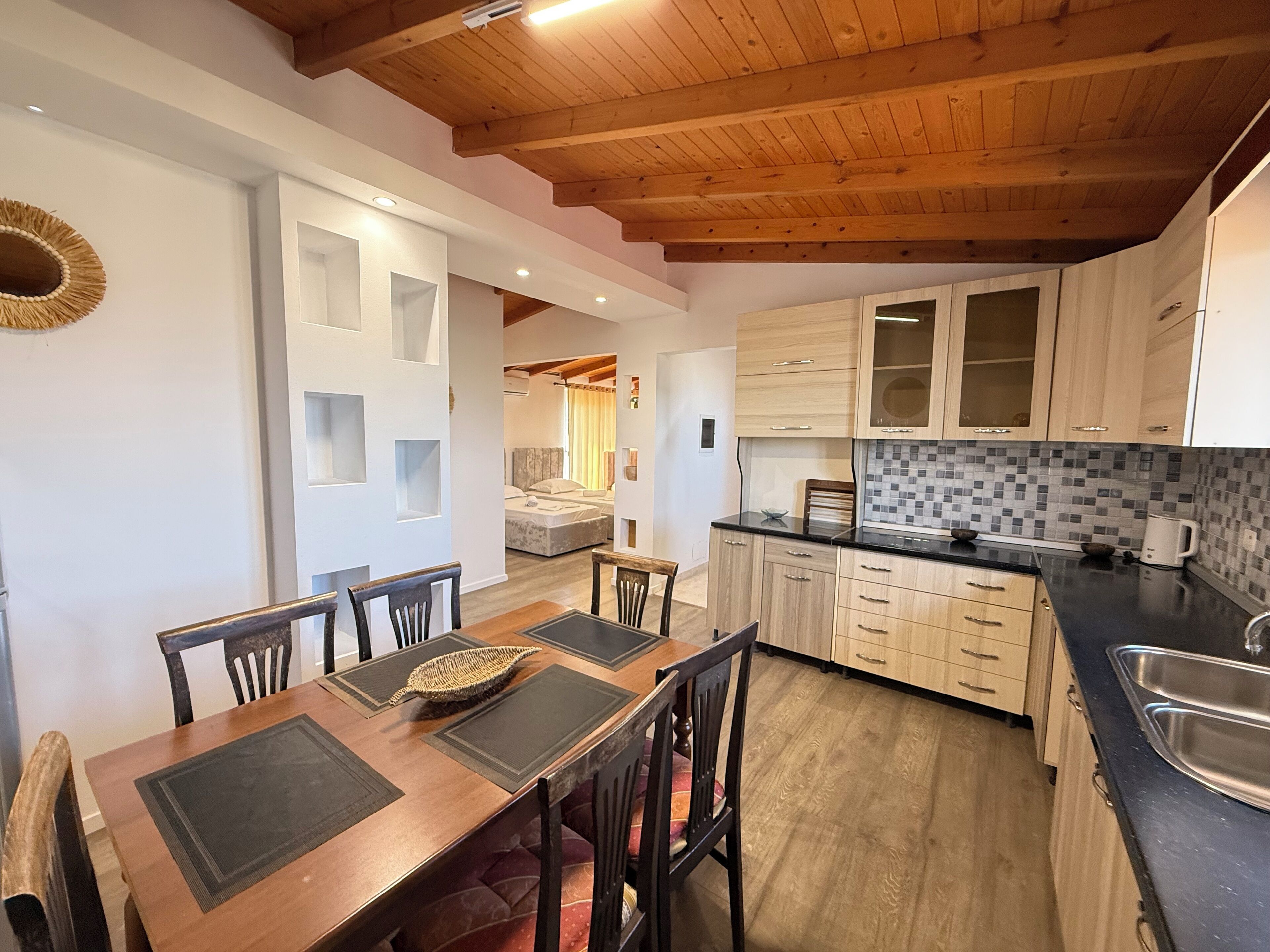 A kitchen with wooden cabinets, a dining table with chairs, and a visible bedroom with a bed.