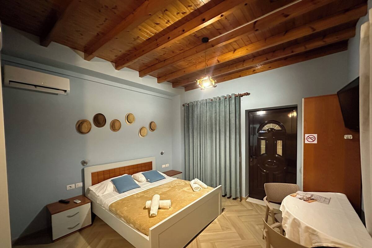 A bedroom with a wooden ceiling, a bed with a white headboard, a nightstand, a chair, a table with a tablecloth, a door with a sign, and a wall with hats.