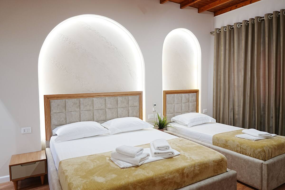 A hotel room with two beds, a nightstand, a plant, and a large arched window.