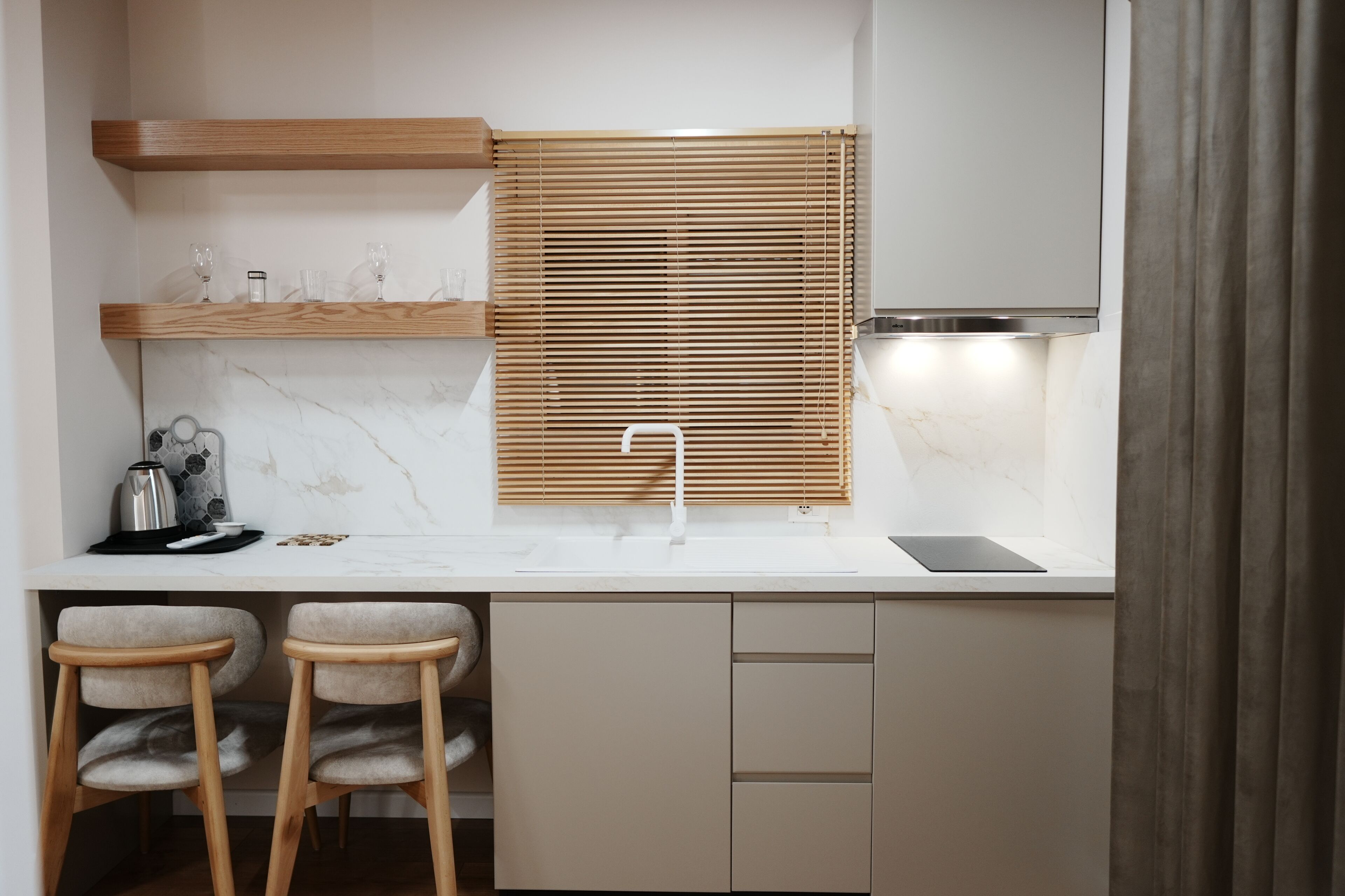 Una cocina moderna con encimera de mármol, persianas de madera, un lavamanos de acero inoxidable y dos taburetes de bar.