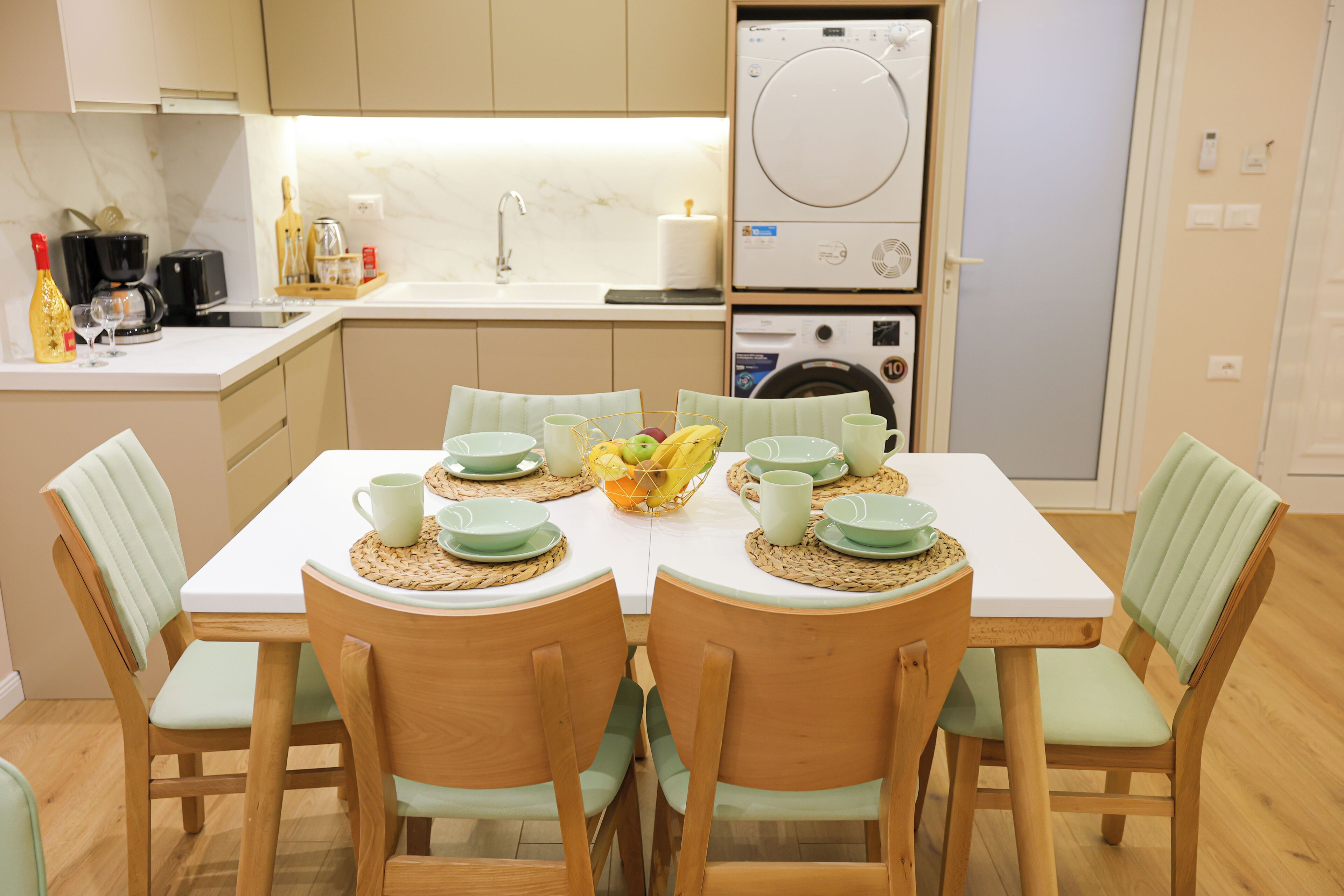 A modern kitchen with a dining table set for four, equipped with a coffee maker, toaster, and washing machine.