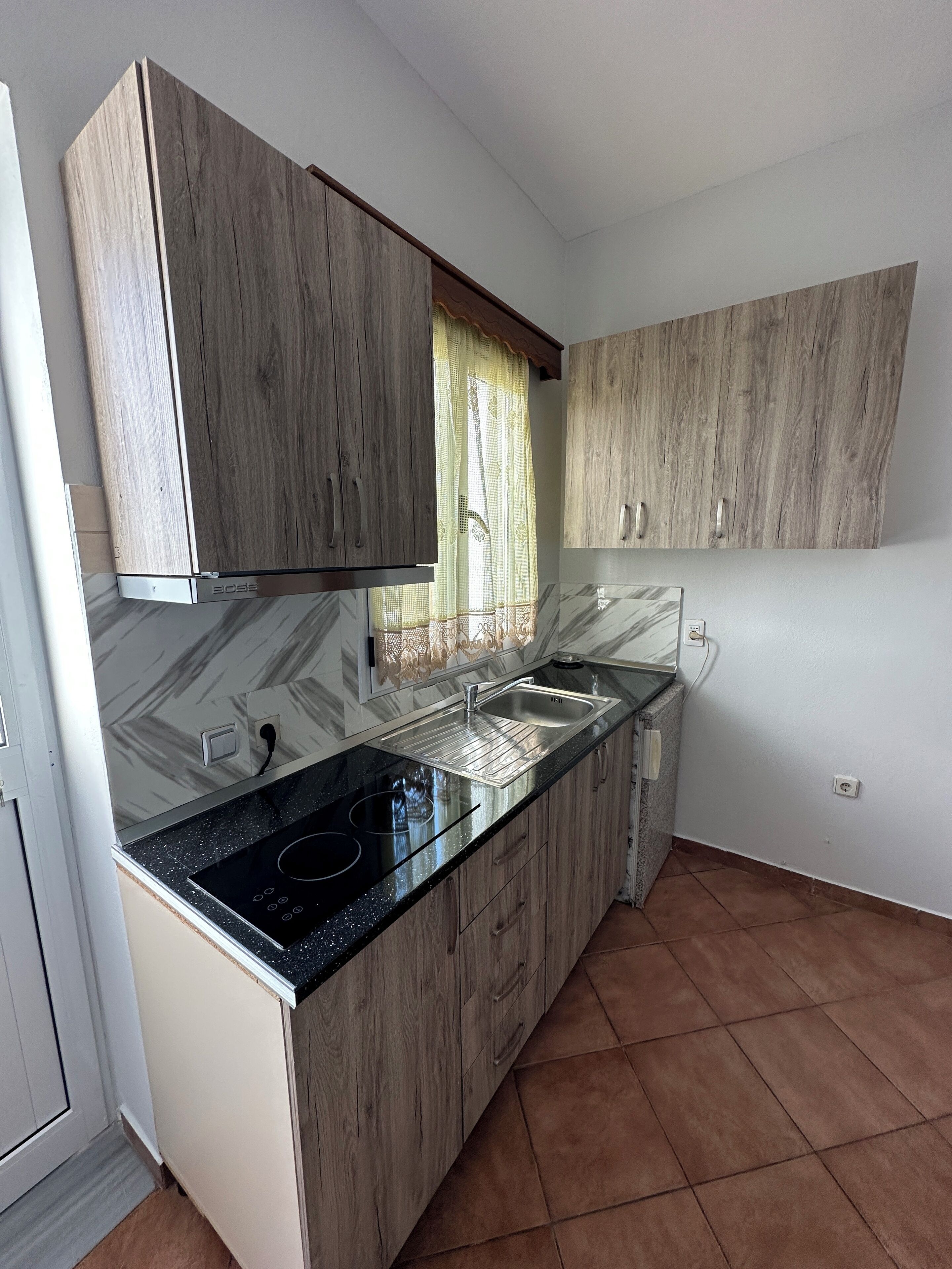 Una cocina con armarios de madera, encimera negra, un lavabo y una ventana con cortina.