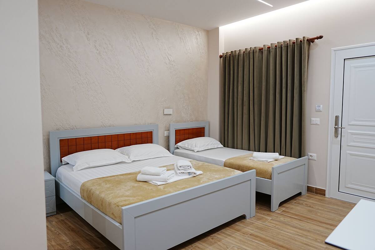 A hotel room with two single beds, a wooden floor, a white door, and a window with curtains.