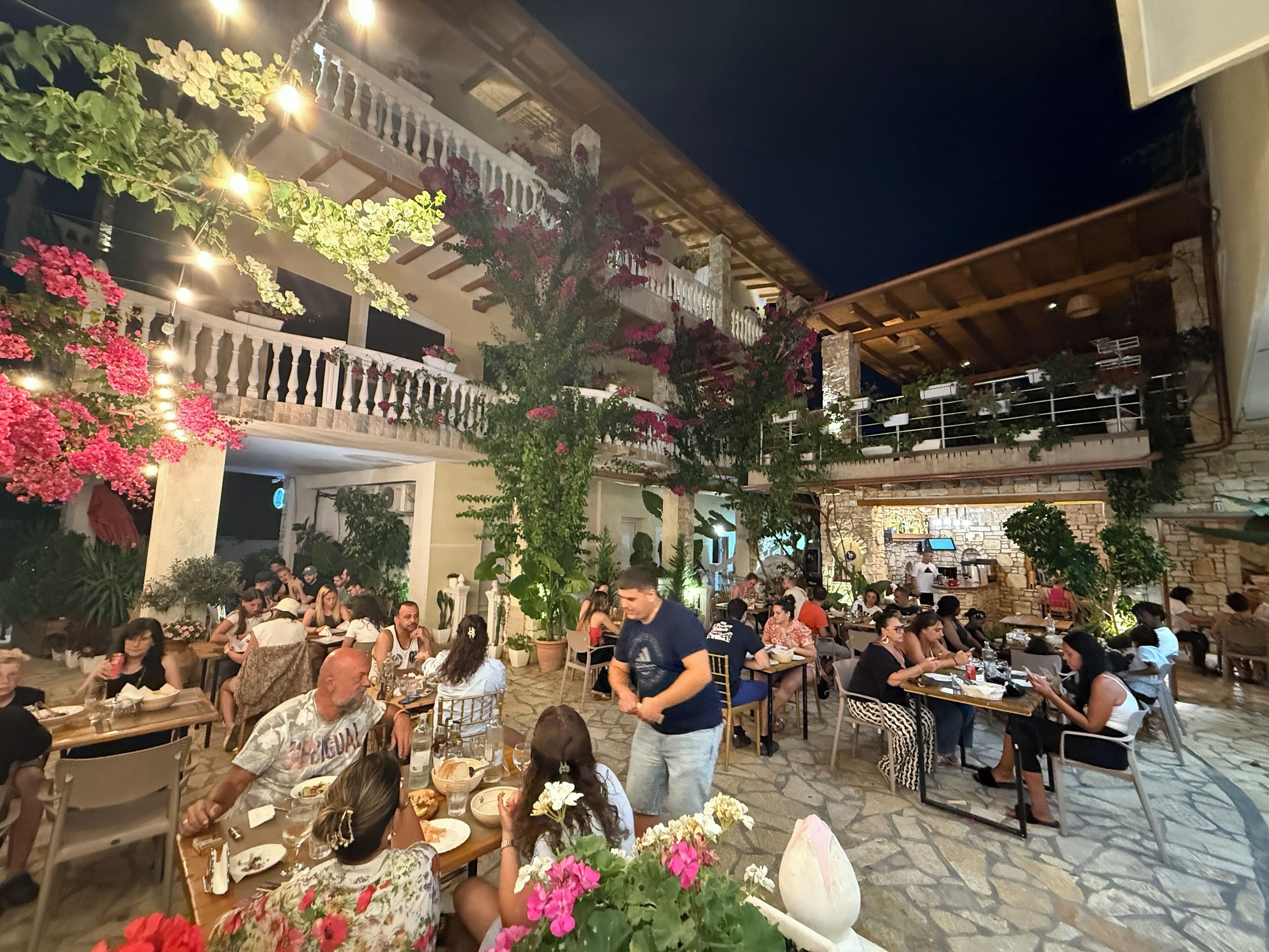 An outdoor dining area with multiple tables and chairs, surrounded by potted plants and flowers.