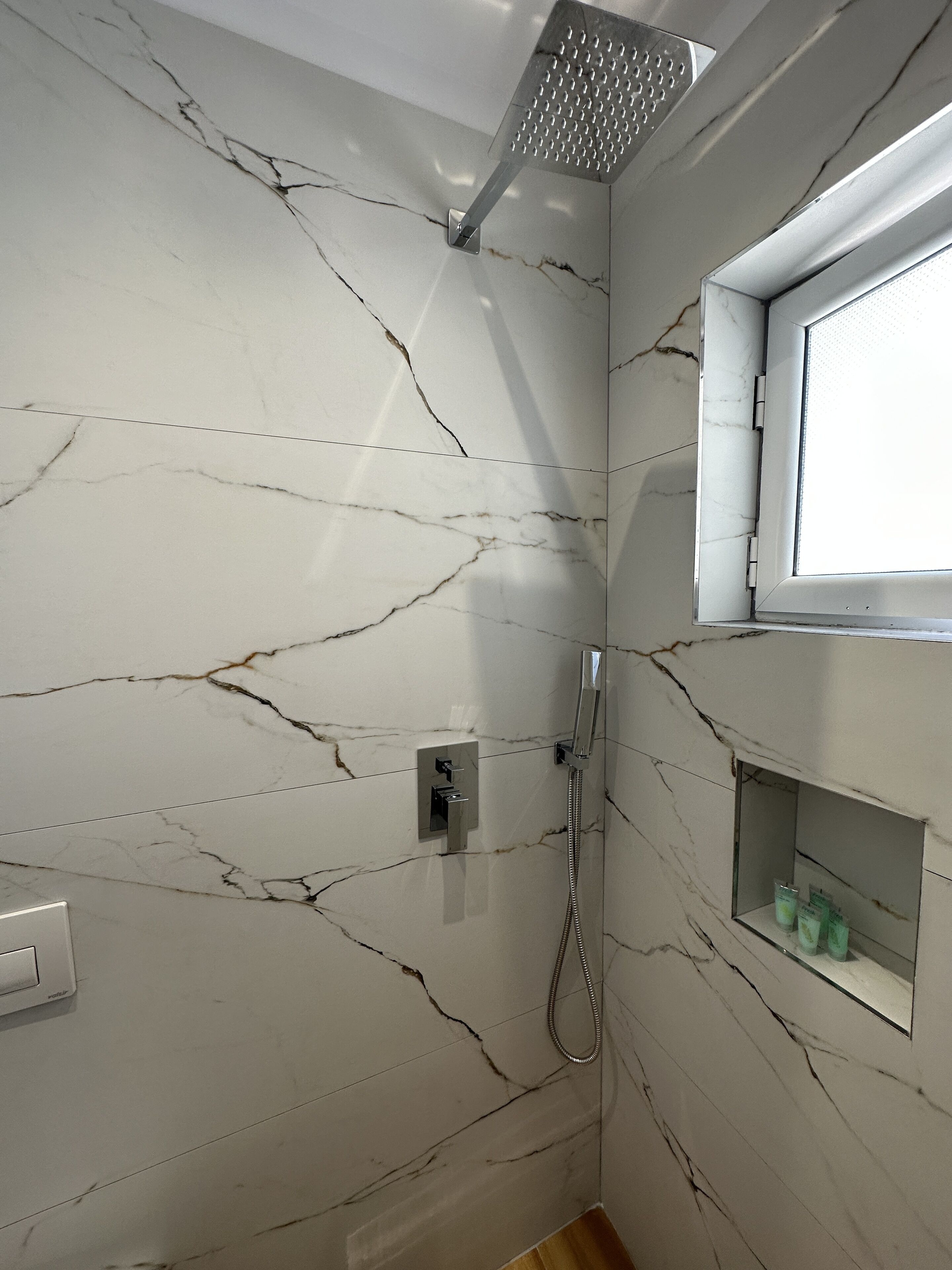 Bagno con pareti in marmo bianco, soffione doccia, doccetta manuale, una finestra e uno scaffale con articoli da toilette.