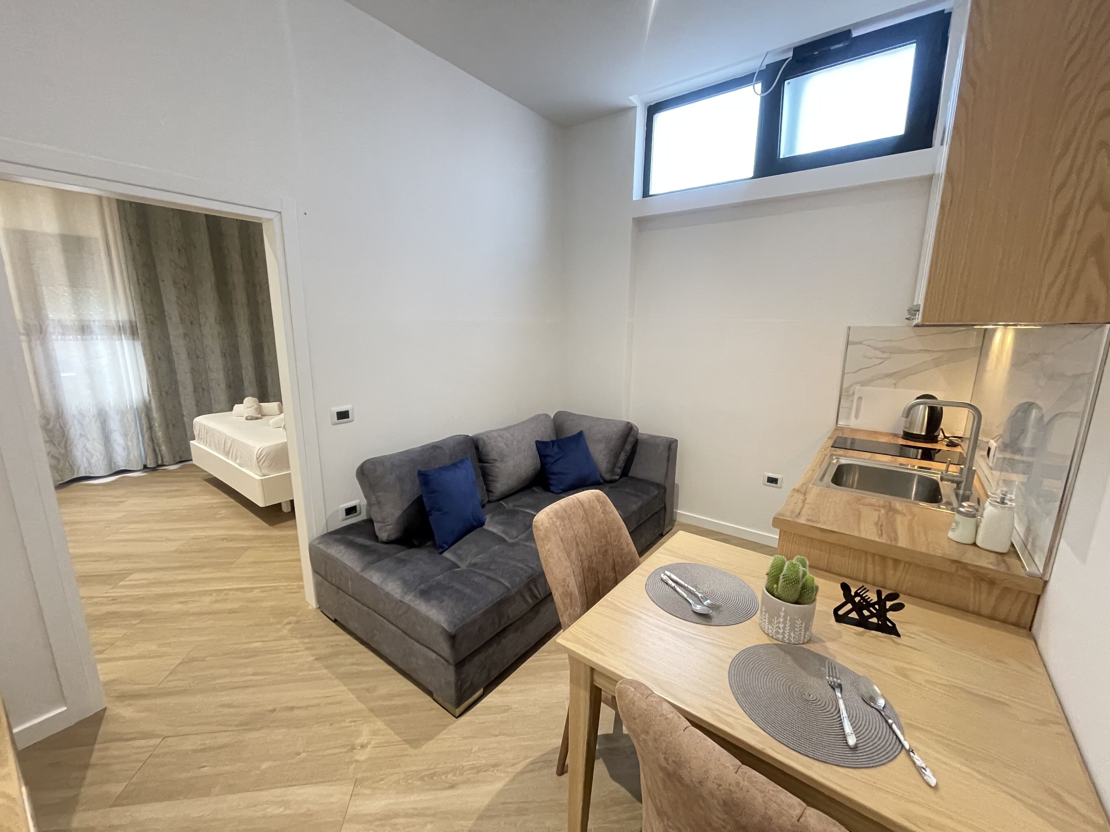 A compact living space with a grey sofa, a dining table with cutlery, a small kitchen area, and a window with natural light.